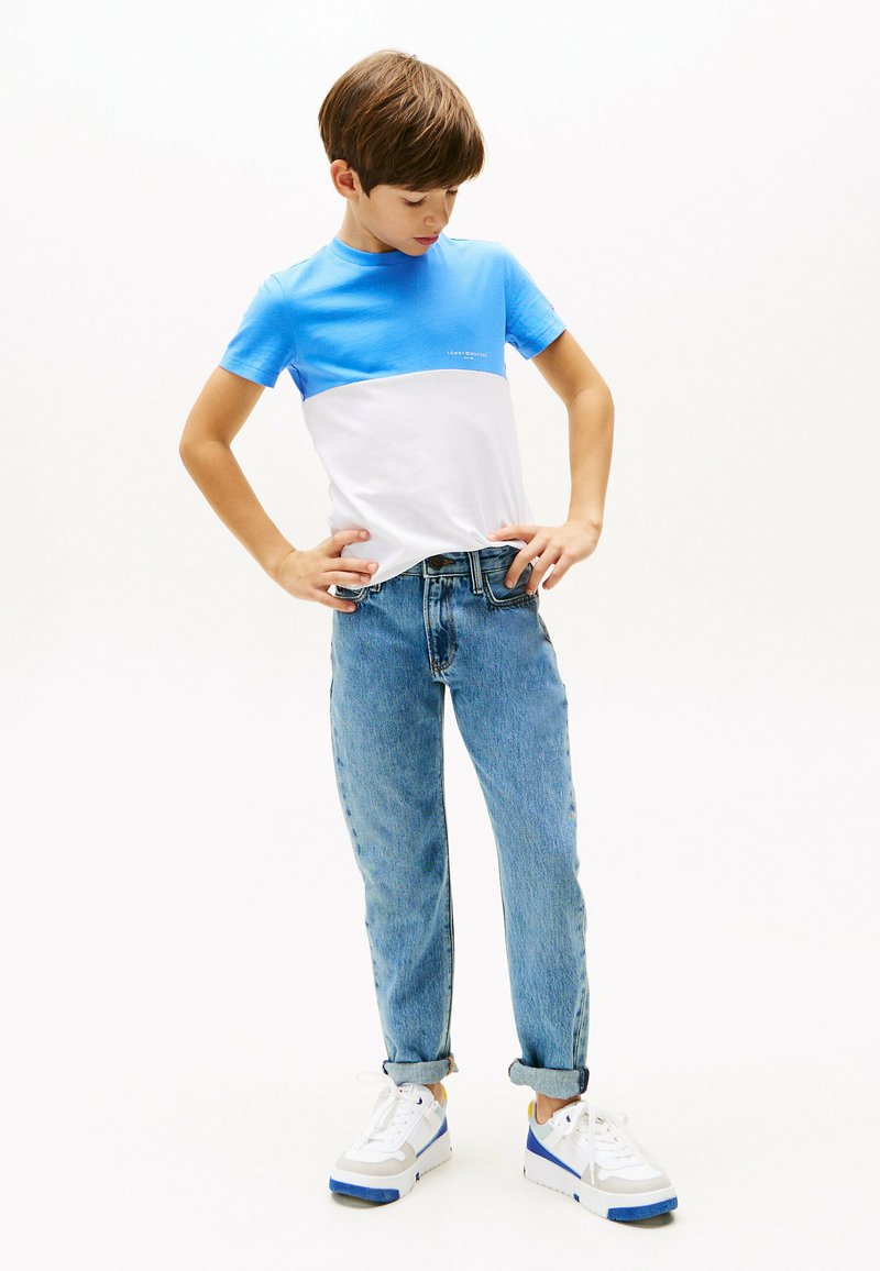 T-shirt con blocchi di colore blu e bianco, maniche corte, collo rotondo. Indossata con jeans in denim chiaro arrotolati e sneakers bianche con accenti blu.