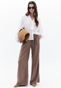 Witte blouse met lange mouwen, gecombineerd met bruine wijde broek en een geweven beige hoed. Zonnebril maakt de outfit compleet.