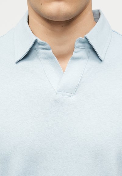 Άνδρας που φοράει ανοιχτό μπλε πουκάμισο με γιακά και ανοιχτό V-λιγότερο, που δείχνει τον λαιμό και το κάτω μέρος του προσώπου.