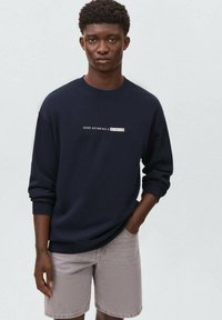 Felpa blu navy con scollo a girocollo, con testo bianco "OGNI AZIONE HA UNA RE/AZIONE" sul petto, abbinata a pantaloncini grigi.