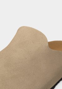 Κοντινό πλάνο ενός μπεζ σουέντ παπουτσιού χωρίς κορδόνια, με στρογγυλεμένη μύτη και λεία υφή, σε απλό φόντο.