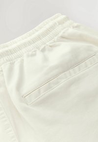 Pantalon de couleur crème avec une taille élastique, une poche arrière unique et une texture lisse tissée.