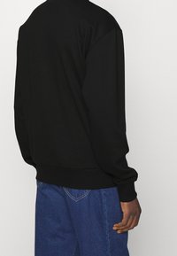 Felpa nera con una vestibilità comoda, polsini e orlo a costine, realizzata in tessuto liscio. Abbinata a jeans blu con un delicato dettaglio di cucitura.
