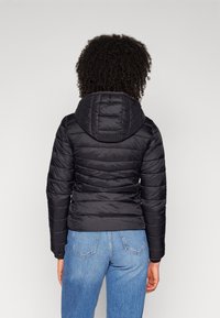 Sort pufferjakke med hætte, prydet med kviltede linjer og en tætsiddende silhuet. Båret over blå jeans, der fremhæver tekstur og design detaljer.