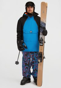Chaqueta de esquí azul y negra con capucha; pantalones vibrantes con estampado en negro y rosa; botas de esquí negras; sosteniendo esquís y bastones; equipado para esquiar.