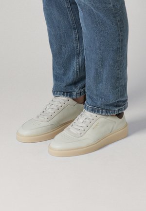 Persona que lleva zapatillas de cuero blancas bajas con suelas beige y jeans de mezclilla azul sobre un fondo liso.