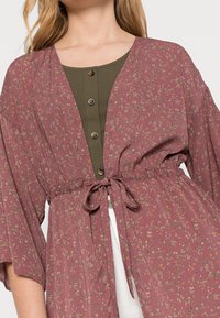 Flowy mauve kimono with floral pattern, wide sleeves, and a tie waist. Underneath, a green button-up top adds contrast to the outfit.