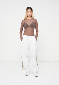 Young woman standing with hands in pockets wearing sheer brown long-sleeve top over black bra, white wide-leg pants, and white sneakers.