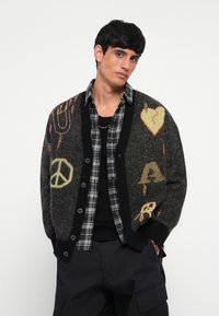 Cárdigan negro de punto con patrones coloridos (corazones, símbolo de paz) sobre una camisa de cuadros gris con botones. Ajustado con un cuello y puños de canalé.