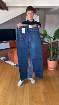 Jovem segurando jeans azuis oversized com etiquetas numa sala de estar com piso de madeira, sofá laranja, TV e uma grande planta em vaso.