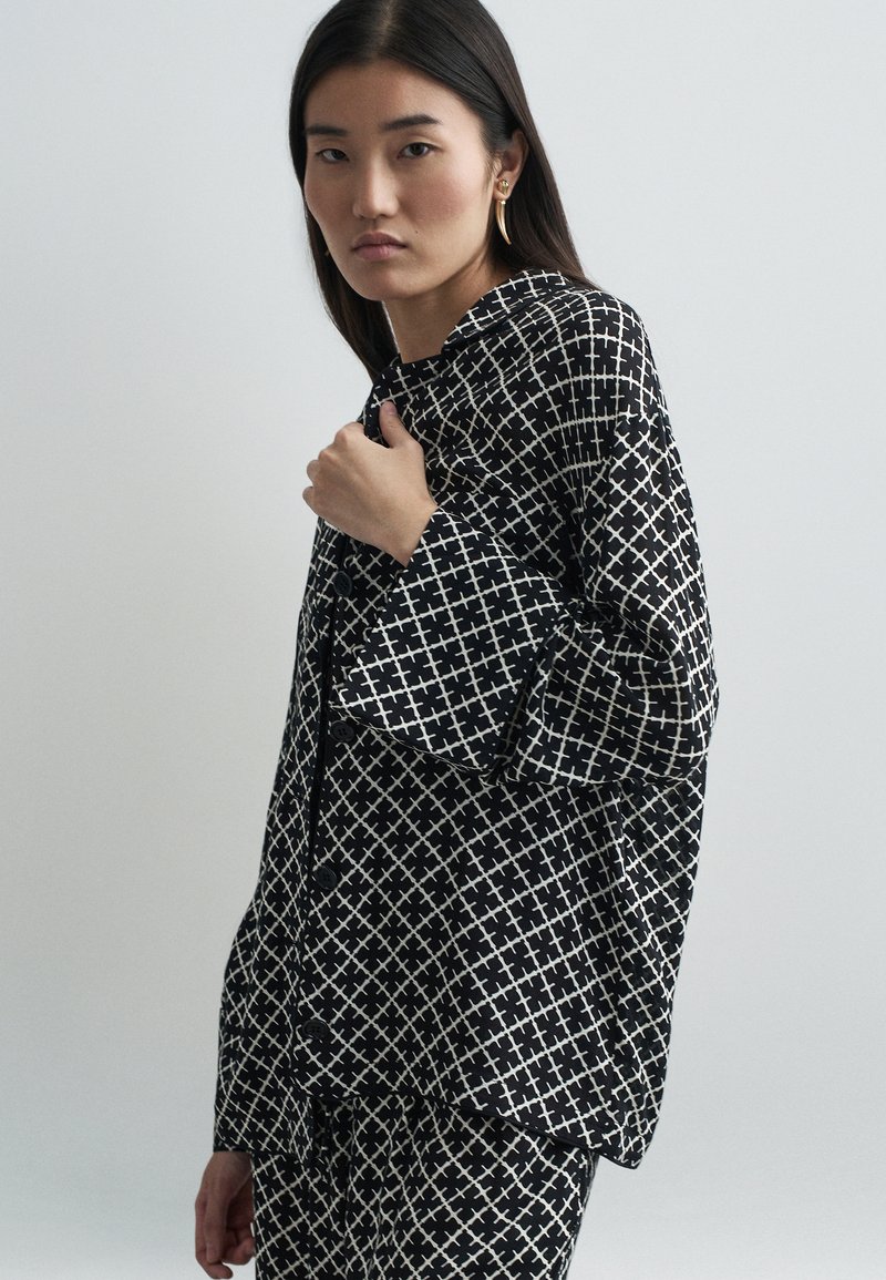 Μαύρο και άσπρο πουκάμισο με σχέδιο γεωμετρικού �σταυρού, διαθέτοντας χαλαρή εφαρμογή και φαρδιά μανίκια, κατασκευασμένο από μαλακό ύφασμα.