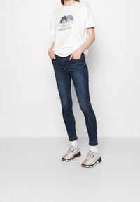 Vit t-shirt med grafiskt tryck, mörkblå tajta jeans med fransad nederkant och beige sportskor med rosa och grå accenter.