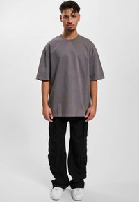 Grå oversized t-shirt med korte ærmer, rund halsudskæring og et diskret logo på venstre bryst; kombineret med sorte cargo-bukser og hvide sneakers.