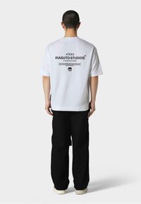 Hvit oversized t-skjorte med svart grafisk tekst på baksiden, parret med løse svarte bukser og beige sko. Minimalistisk design, uten mønstre.