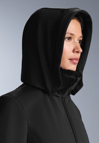 Crni kaput s kapuljačom i mekim, krznenim ovratnikom. Glatka tkanina, krojena fit s vidljivim šavovima. Kapuljača je velika i nosi se podignuta, naglašavajući pokrivenost.