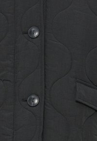 Veste noire matelassée avec un tissu texturé, des boutons ronds noirs et des motifs en vagues cousus sur le devant et les accents des manches.