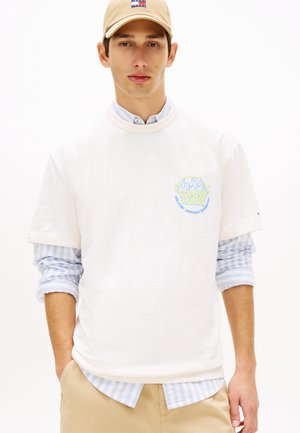 Νεαρός άνδρας που φοράει μπεζ καπέλο, λευκό T-shirt Tommy Jeans πάνω από μπλε ριγέ πουκάμισο και μπεζ παντελόνι με τα χέρια στις τσέπες.