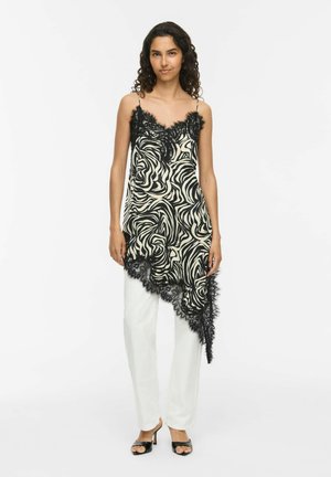Femme portant un caraco asymétrique à motif abstrait noir et blanc avec bordure en dentelle, sur un pantalon blanc et des talons ouverts noirs, debout sur fond blanc.