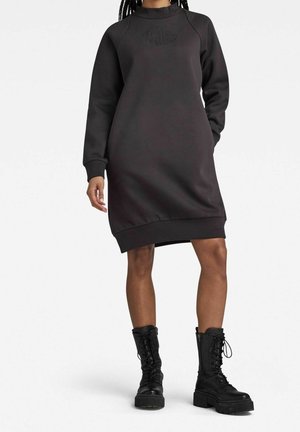 Person iført en sort sweatshirtkjole med lange ærmer og ribbede manchetter samt sorte snørebånds-kampstøvler, stående mod en ensfarvet baggrund.