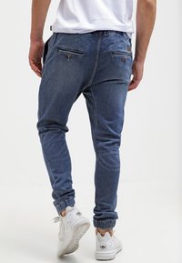 Modré denimové jogger kalhoty s elastickými manžetami, se dvěma zadními kapsami na knoflíky, oblečené s bílými teniskami.