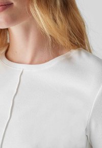 Weißes, geripptes Oberteil aus Stoff mit rundem Ausschnitt, das eine zentrale Nahtdetails aufweist. Die Textur wirkt weich und leicht dehnbar.