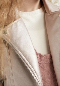 Chaqueta de cuero rosa metálico con un diseño ajustado; presenta cremallera plateada y detalles de botones a presión. Debajo, una blusa texturizada de color rosa claro.