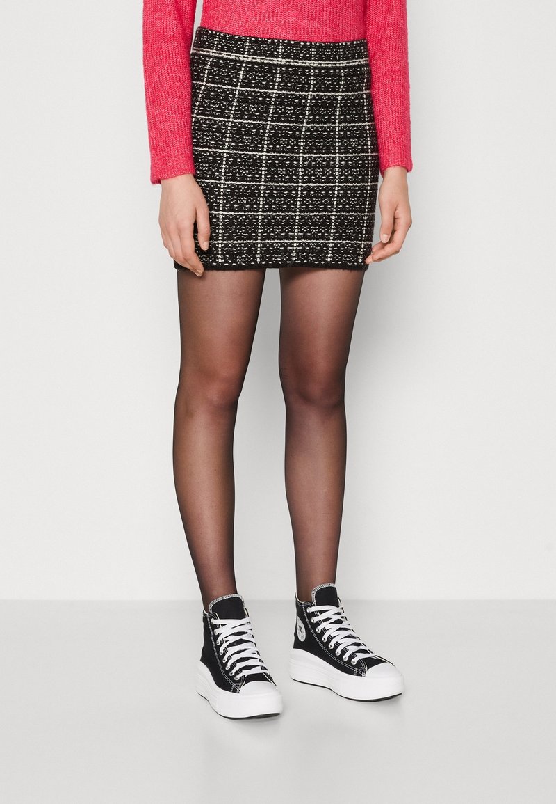 Femme portant une jupe à carreaux noir et blanc, des collants noirs transparents, des baskets montantes noires et un pull rouge à manches longues.