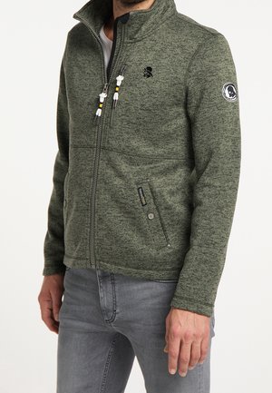 Homme portant une veste polaire verte zippée avec des poches avant zippées et un patch logo noir sur le haut du bras et la poitrine.