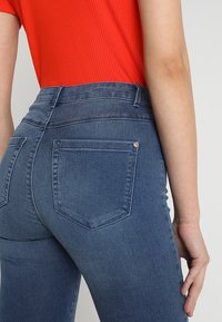 High-waisted, blue denim jeans with a smooth texture and two back pockets. The design includes a small metal accent at the pocket.