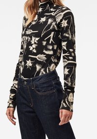 Langærmet sort top med cremefarvet botanisk print; tætsiddende design, høj hals, blødt stof. Kombineret med mørk denim jeans.