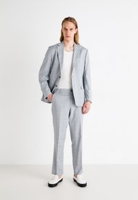 Completo grigio chiaro con finitura testurizzata, caratterizzato da rever a punte, chiusura a due bottoni e pantaloni dritti. Abbinato a un top bianco e scarpe.