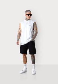 White sleeveless shirt with logo, black shorts, white sneakers, and patterned tattoos. The model wears dark sunglasses.