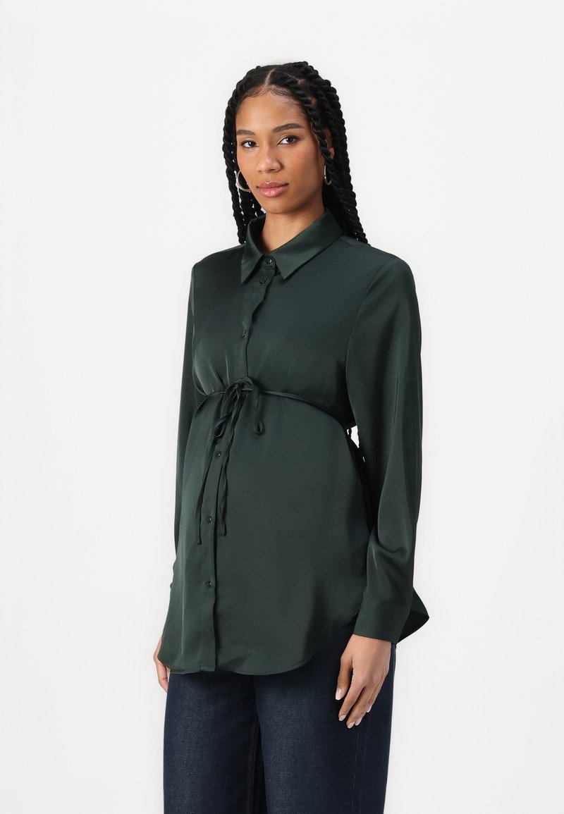 Σκούρο πράσινο πουκάμισο με σατινέ υφή, με γιακά, κλείσιμο με κουμπιά μπροστά και λεπτομέρεια με γραβάτα κάτω από το στήθος. Μακριά μανίκια και χαλαρή εφαρμογή.