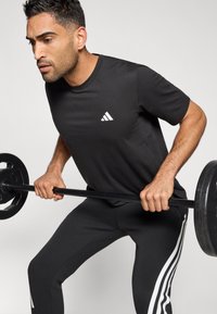 Juodas sportinis marškinėlis su balta Adidas logotipu, derinamas su juodomis treniruočių kelnėmis, turinčiomis baltas juosteles, dėvimas svorį kilnojant.