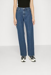 Jeans largos azuis feitos de denim, com cintura alta, design de cinco bolsos e corte reto, combinados com sapatilhas pretas e brancas.
