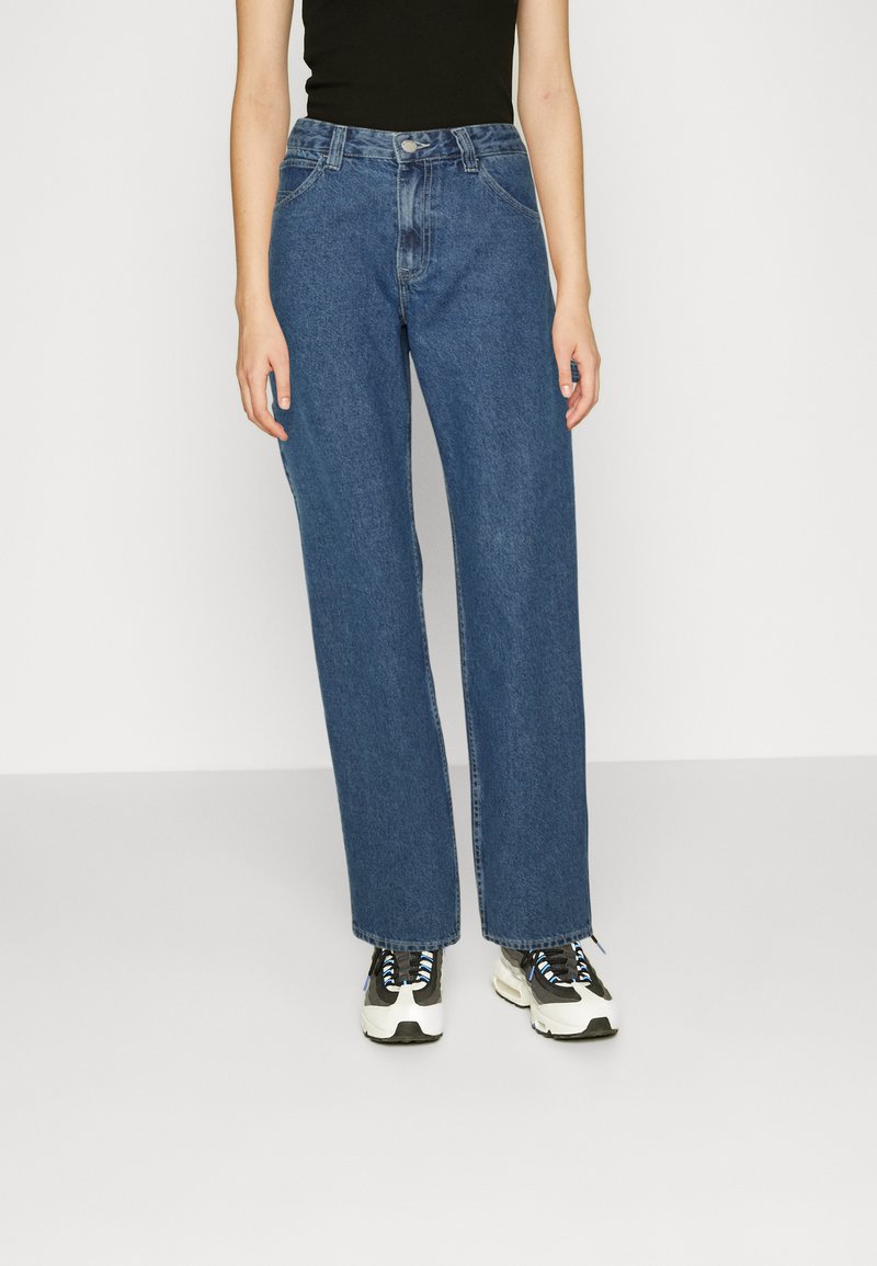Jeans largos azuis feitos de denim, com cintura alta, design de cinco bolsos e corte reto, combinados com sapatilhas pretas e brancas.