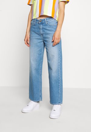 Persona vestida con vaqueros anchos de pierna en azul claro, un top corto de rayas multicolores y zapatillas blancas, de pie sobre un fondo liso.