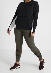 Haut noir à manches longues avec panneaux latéraux blancs, associé à des leggings vert olive avec découpes en mesh et chaussures de sport noires.