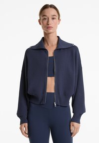 Marineblaue strukturierte Zip-Jacke mit breiter Kragen und langen Ärmeln, ausgestattet mit einem gerippten Saum und subtilen weißen Akzenten am Kragen.