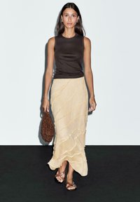 Haut sans manches marron, jupe maxi beige texturée avec des lignes ondulées, sac en perles marron et sandales plates marron. Fond uni.