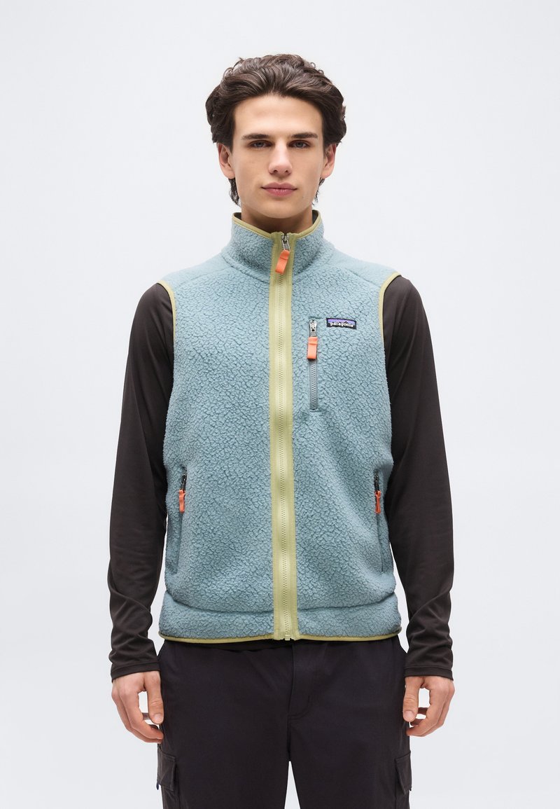 Νέος άνδρας που φοράει ανοιχτό μπλε φλις γιλέκο με φερμουάρ σε πορτοκαλί χρώμα πάνω από μαύρο μακρυμάνικο πουκάμισο, στέκεται μπροστά σε απλή φόντο.