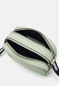 Zielona skórzana torebka na ramię z dwoma zamykanymi na zamek przegródkami, czarnym wnętrzem i odpinanym paskiem z białymi akcentami.