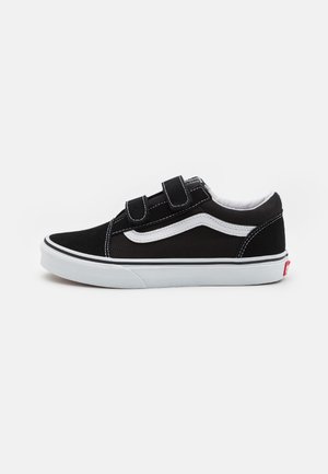 Baskets basses noires avec une bande blanche, une semelle blanche et deux brides velcro noires sur fond clair uni.