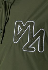 Olivenfarbener Stoff mit glatter Textur, der ein auffälliges, weißes und dunkelgrünes gesticktes Logodesign auf der Brust aufweist.