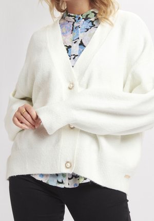 Kvinde iført en stor hvid cardigan med perleknapper over en blomsterbluse og sorte bukser, med hånd der justerer ærmet.