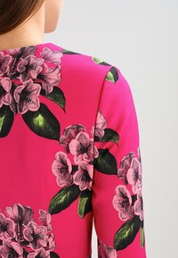Fuchsia Kleid mit floralem Muster, das rosa und schwarze Blumen zeigt, akzentuiert durch grüne Blätter. Der Stoff hat eine glatte, leicht glänzende Textur.