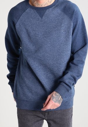Homme portant un sweat-shirt à manches longues bleu marine et un jean foncé, avec des tatouages visibles au cou et aux mains, debout devant un fond uni.