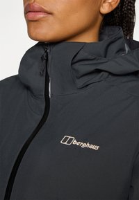 Nærbillede af en person iført en sort jakke med "Berghaus"-logoet på siden, der viser kraven og lynlåsdetaljerne.