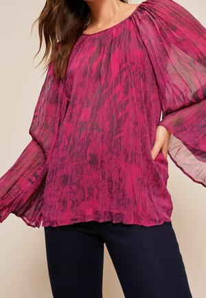 Eine Frau trägt eine durchsichtige, langärmelige, plissierte fuchsienfarbene Bluse mit floralem Muster und weiten, fließenden Ärmeln, kombiniert mit dunkler Hose.