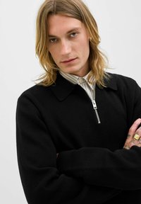 Blondýna s dlouhými vlasy, oblečená v černé svetr s zipem přes pruhovanou košili, s překříženýma rukama, ukazující zlatý prsten a tetování.